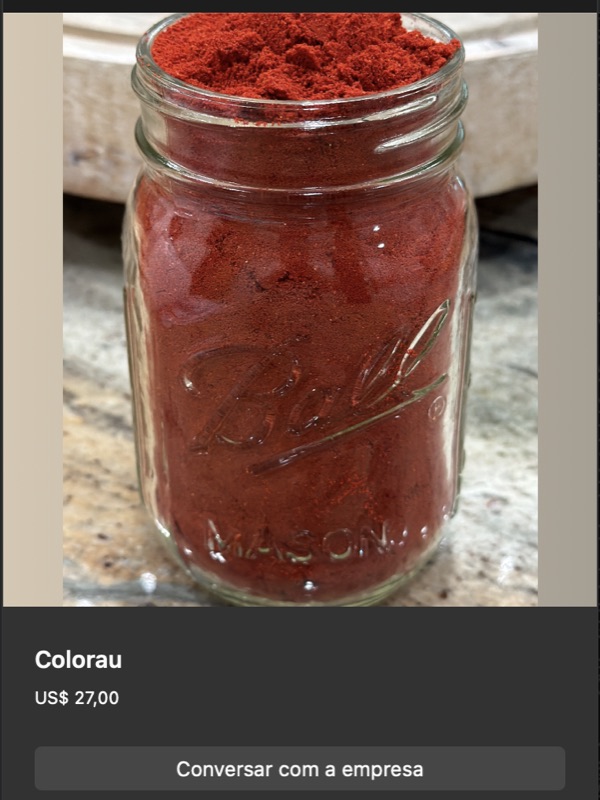 Pote de vidro tipo Ball mason jar cheio de colorau (urucum) em pó vermelho intenso. Pequeno monte de pó saindo da boca do pote, evidenciando textura fina. Iluminação destaca o pigmento avermelhado.