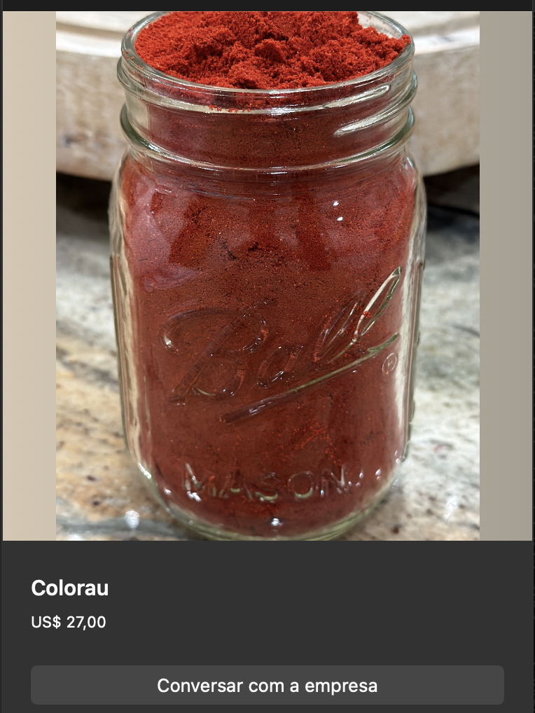 Pote de vidro tipo Ball mason jar cheio de colorau (urucum) em pó vermelho intenso. Pequeno monte de pó saindo da boca do pote, evidenciando textura fina. Iluminação destaca o pigmento avermelhado.